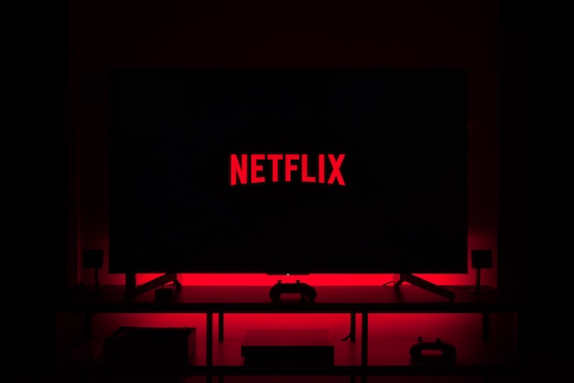 Telewizor z wyświetlonym logiem "Netflix" na czarnym tle. Przed monitorem czerwone krzesła