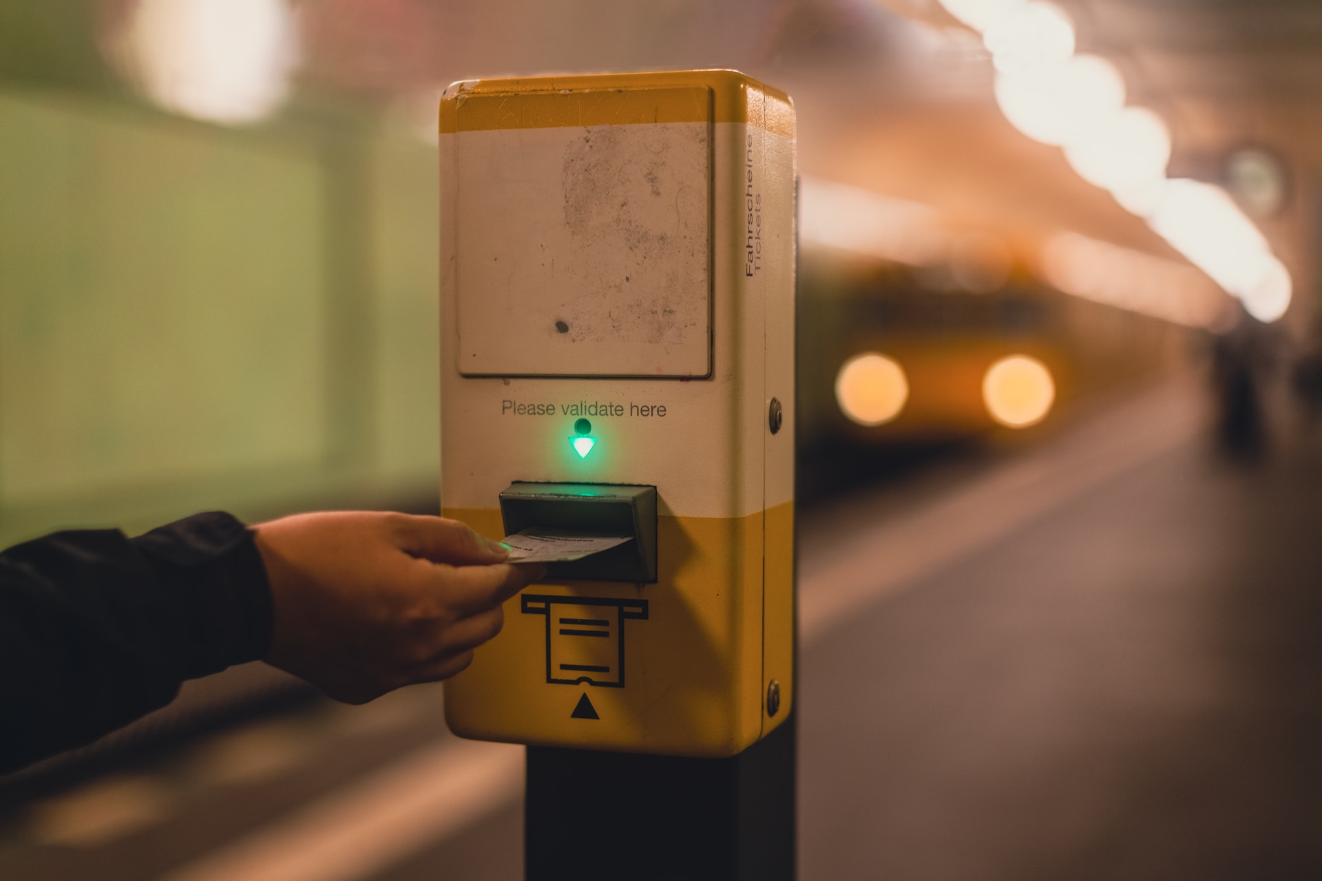 Osoba wkłada bilet do elektronicznego kasownika. W tle nadjeżdżający pociąg metra
